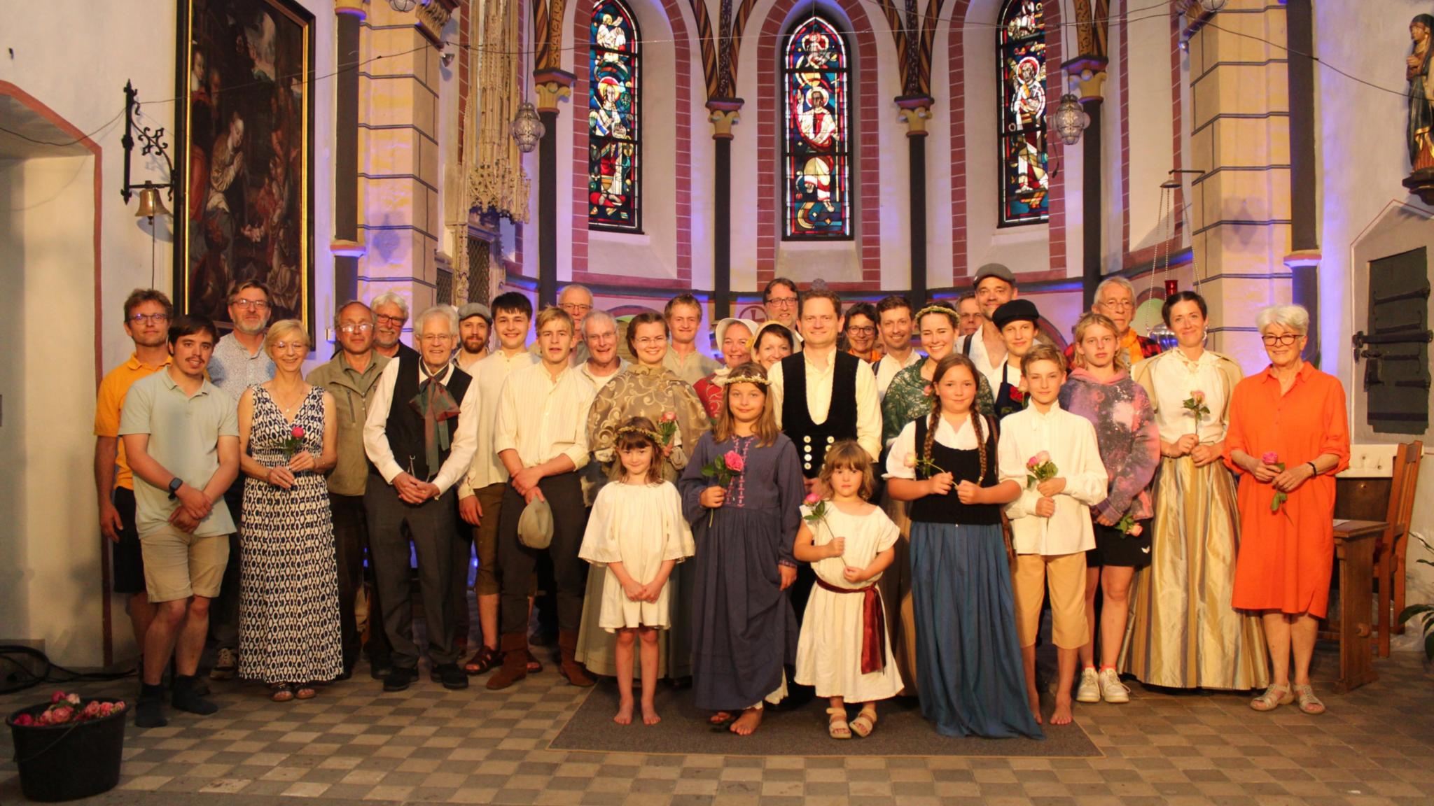 Eine große Gruppe von Menschen steht gemeinsam im Altarraum einer Kirche. Viele tragen historische oder festliche Kleidung, einige moderne Kleidung. Kinder und Erwachsene stehen gemischt in mehreren Reihen. Hinter ihnen sind hohe, bunt leuchtende Kirchenfenster und gotische Architektur zu erkennen.